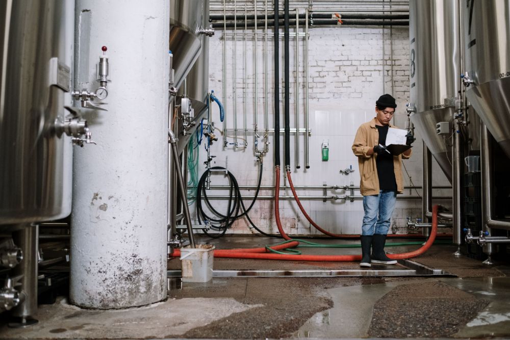 An employee with a clipboard inspects tanks and pipes in a production hall
