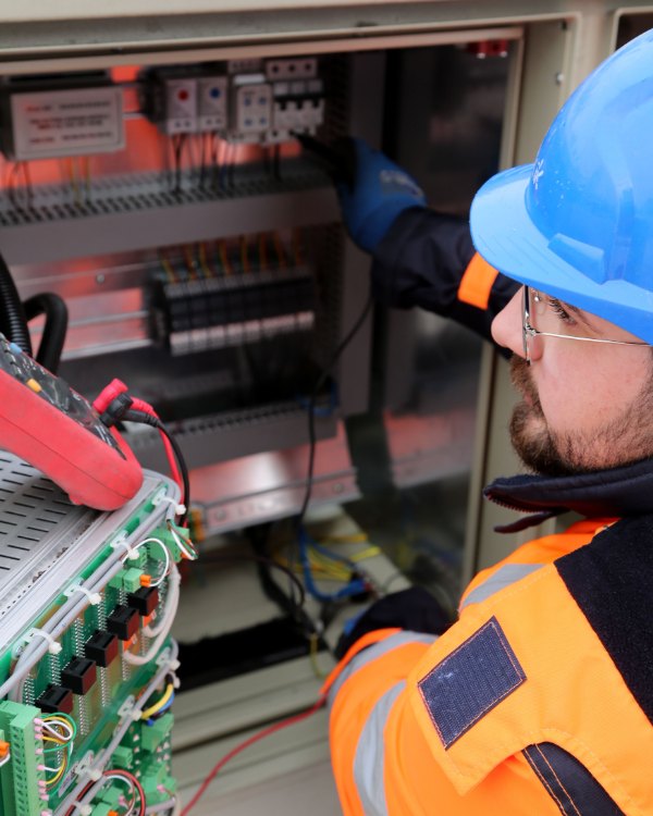 An electrician in a hard hat and high-visibility jacket tests a switch cabinet with a meter